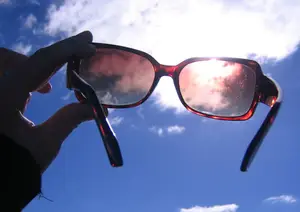 A pair of red sunglasses with brown lenses being held up in the sky with clouds in the background.