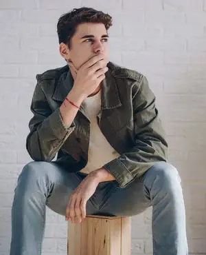 Man sitting on a wooden stool wearing a green jacket and blue jeans with his hand on his chin.