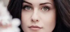 Close-up of a woman's face with long dark hair, green eyes, and a thoughtful expression, holding a white object in her hand.