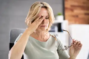 A woman sitting at a desk and rubbing her temples with her eyes closed, holding a pair of glasses in her other hand