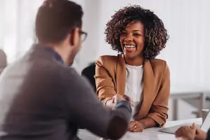 A smiling woman with curly hair shakes hands with a man wearing glasses in a meeting room