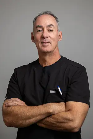 A man wearing a black shirt with a tag on the left side and a pen on the right side, standing in front of a gray background with his arms crossed and smiling at the camera