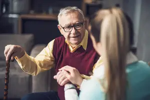 An elderly man holding a cane and sitting on a couch with a woman in front of him