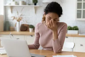A woman with her hand covering her face while sitting at a desk with a laptop in front of her.