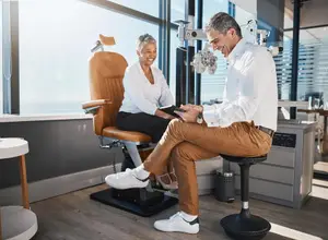 A man and a woman are sitting in a chair in front of a window in an office. The man is wearing a white long-sleeve shirt and pants. The woman is wearing a white blouse and pants. The man is looking at the woman while smiling. There is a table and a desk with drawers on the right side. There is a trash bin and a cabinet on the left side.