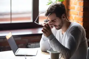 A man sitting at a desk is rubbing his eyes and has his hand on his forehead. He is wearing a white shirt and has a laptop, a cup, and a pen on the desk. Behind him is a window with a potted plant and a brick wall.