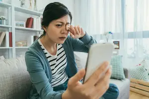 A woman is sitting on the couch, looking at her phone, and rubbing her nose with her hand.