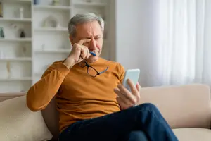An older man with gray hair wearing glasses and an orange shirt is sitting on a couch while holding a phone and a pen, and he seems to be in pain.