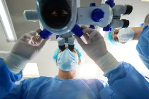 Three surgeons in blue gowns and masks are performing surgery in a well-lit operating room, using a microscope.