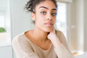 A woman with curly hair wearing a beige sweater is sitting in a room and looking forward with her right hand on her chin