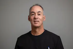 A man wearing a black shirt is smiling and posing for a picture with a gray background.