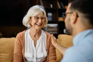 Elderly woman with glasses sitting on a couch smiling at a man who is touching her shoulder