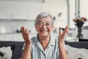 A smiling elderly woman sitting on a couch in a kitchen, wearing glasses and a necklace, with a white blanket on her lap and a vase of flowers on the counter behind her.