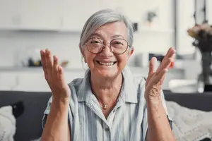 A smiling elderly woman with glasses and a necklace is raising both hands in a room