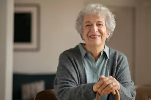 An elderly woman with gray hair smiles and looks forward while standing in a room with a couch and a picture frame on the wall behind her.