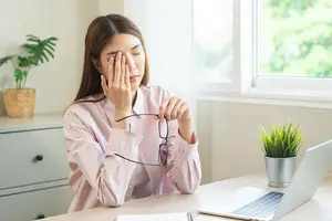 A woman with her eyes closed holding her forehead while sitting in front of a desk