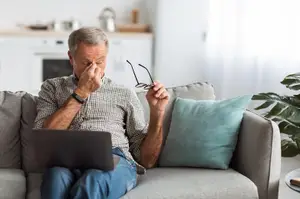 A senior man is sitting on a couch and rubbing his eyes as he suffers from Dry Eye Syndrome