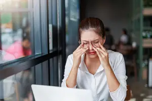 A woman in glasses is holding her forehead with her left hand and looking at something on a laptop in front of her in a coffee shop