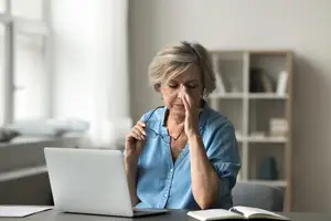 An older woman sitting at a desk with her hand on her forehead looking at a laptop