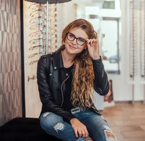 A woman wearing glasses and a leather jacket is smiling while sitting on a bench in a store.