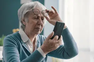 An older woman wearing glasses is holding a smartphone and adjusting her glasses.