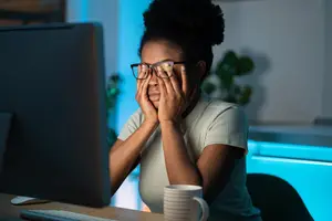 A woman is sitting in front of a computer monitor, covering her eyes with her hands, possibly in a dimly lit room.