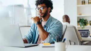A man in glasses is sitting on a chair in front of a desk, typing on a laptop, with a plant and shelves in the background.