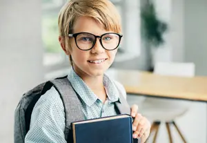 A boy with glasses and a backpack holding a book and smiling
