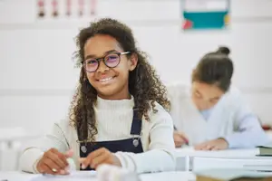 Girl in white turtleneck sweater and glasses smiling while sitting at a desk in a classroom