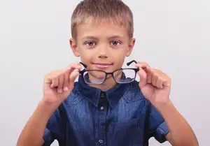 A young boy is holding a pair of glasses in front of his face and smiling.