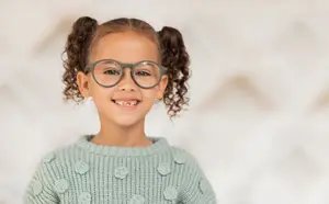 A smiling little girl with curly hair wearing glasses and a green sweater with flower designs.