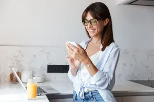 A woman in a kitchen is smiling and looking at her phone.
