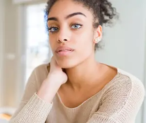 A woman with curly hair and a beige sweater is looking to the side, possibly posing for a photo in a room with a glass window behind her