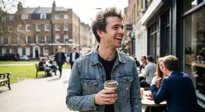 A man holding a coffee cup smiles on a sidewalk with a blurry background of people sitting in cafes and buildings
