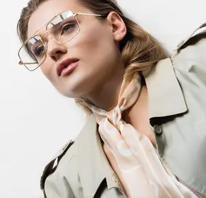 A woman is wearing a beige trench coat, a white scarf, and a pair of gold-framed glasses with clear lenses. She is probably posing for a photo with a white wall in the background.