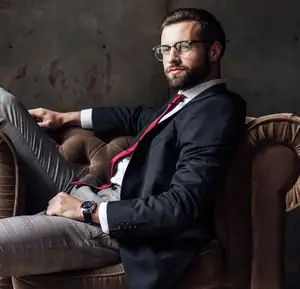 A man sitting on a couch wearing a black suit, glasses, a red tie, and a watch.