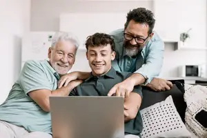 Three people are sitting on a couch looking at a laptop together. One of them is an older man with white hair and a beard, and the other is a young man with a beard and glasses. The third person is a younger man who is smiling and pointing at the laptop. They are all sitting on a couch with pillows. Behind them is a shelf with a plant on it and a microwave.