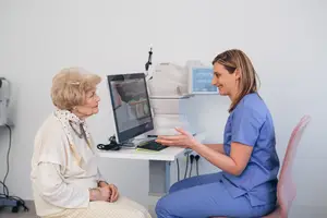 A woman is sitting in front of a computer while another woman wearing blue is smiling beside her.