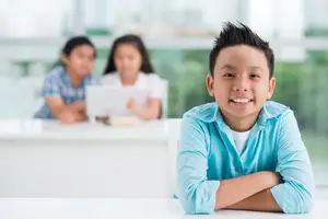 A smiling boy with his arms crossed sitting at a table in front of two girls in a classroom