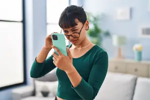 A woman in glasses holding a phone, possibly taking a selfie, standing in a room with a couch, lamp, and plants in the background.