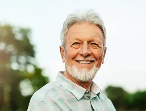 An older man with white hair and a beard is smiling and posing for a picture outdoors in a field.