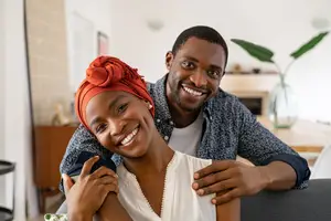 A smiling couple sitting on a couch in a cozy living room with a plant and a wooden table in the background.