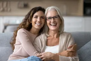 Two women sitting on a couch, one smiling and the other wearing glasses, with their arms around each other
