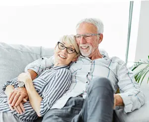 An older couple is sitting on a couch and smiling, probably inside a room with a glass window. The woman is wearing a striped shirt, while the man is wearing a white long-sleeve shirt and glasses. The woman is holding the man's hand, while the man is holding the woman's waist. There is a potted plant on the right side of the couch.