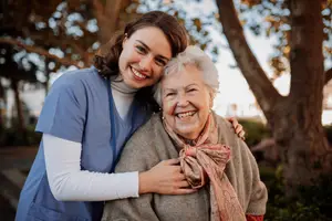 Elderly woman and a young woman wearing blue uniforms and smiling while standing in a garden