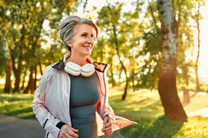 An elderly woman is running on a pathway in a park with trees and grass in the background, and she is wearing a pink jacket, a gray top, and a white neckband with a white earphone.