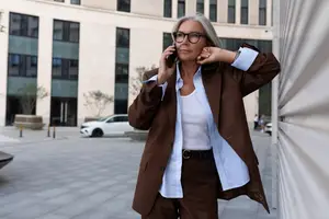 An older woman wearing a suit and glasses is talking on her cell phone in front of a building.