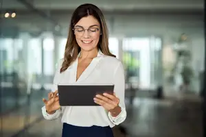 A smiling woman with glasses holding a tablet in an office