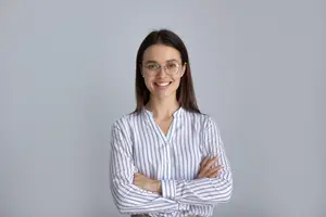 A smiling woman with glasses and a striped shirt standing in front of a gray wall.