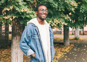 A man in a denim jacket with a white hoodie is smiling and posing for a photo in front of trees in an outdoor area.